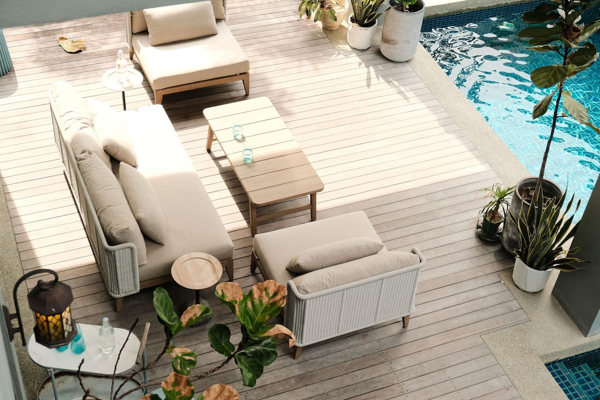 A wooden deck with outdoor furniture, including a sofa, a small table, and a chair. There are potted plants and a pool visible in the background.