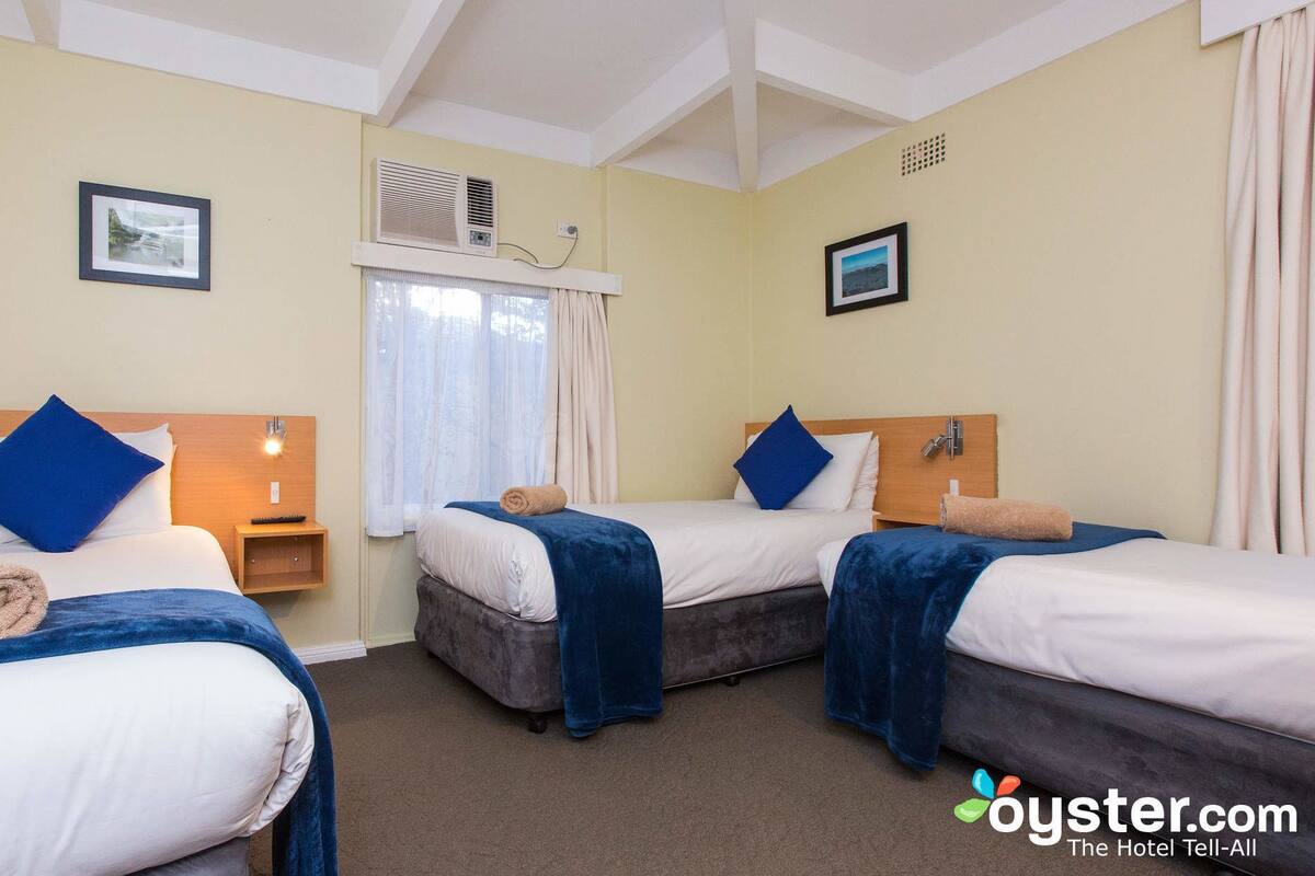 A hotel room with two beds, each with a blue blanket and pillows, a small bedside table with a lamp, and a window with curtains.
