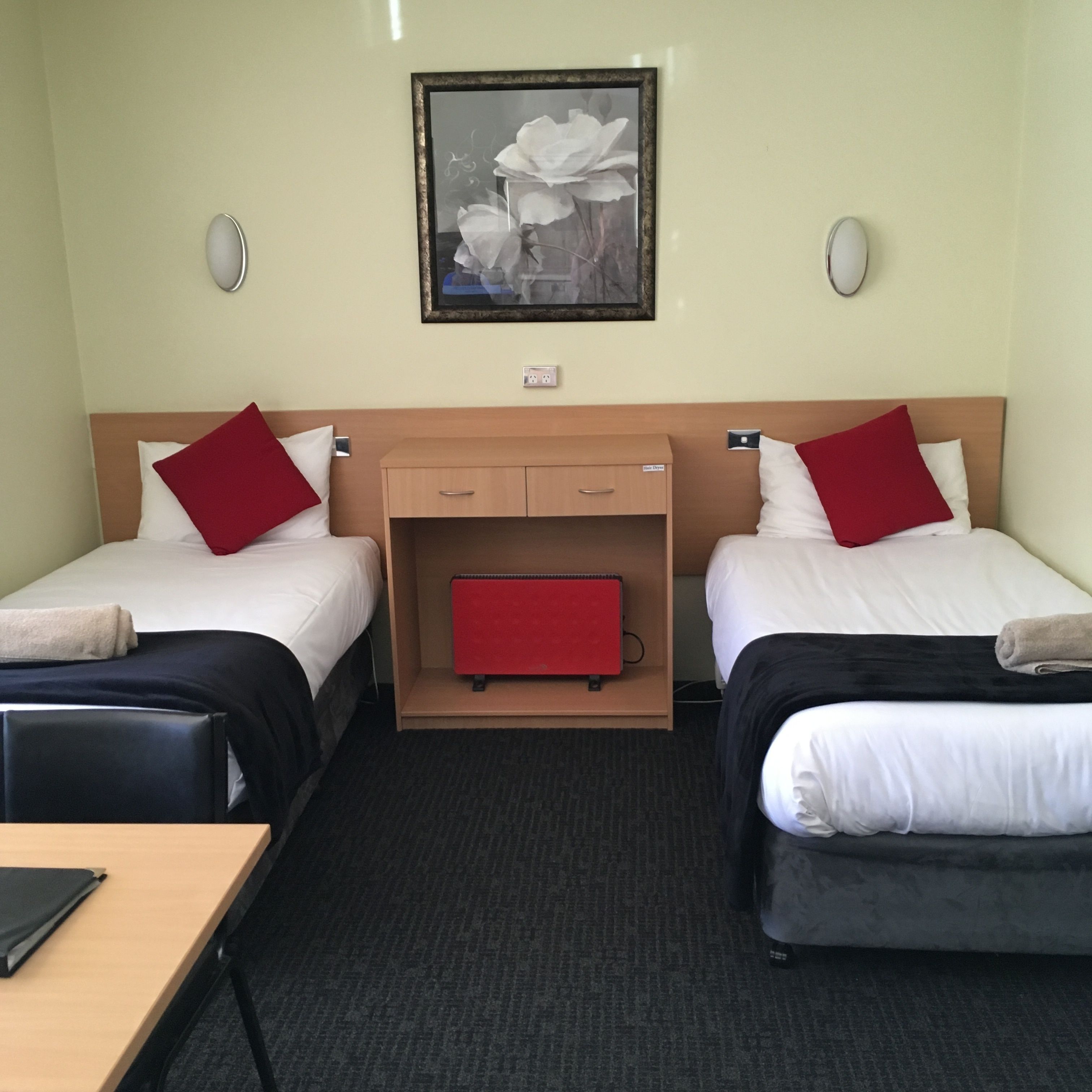 A hotel room with two beds, a small desk, a red suitcase, and a framed picture on the wall.