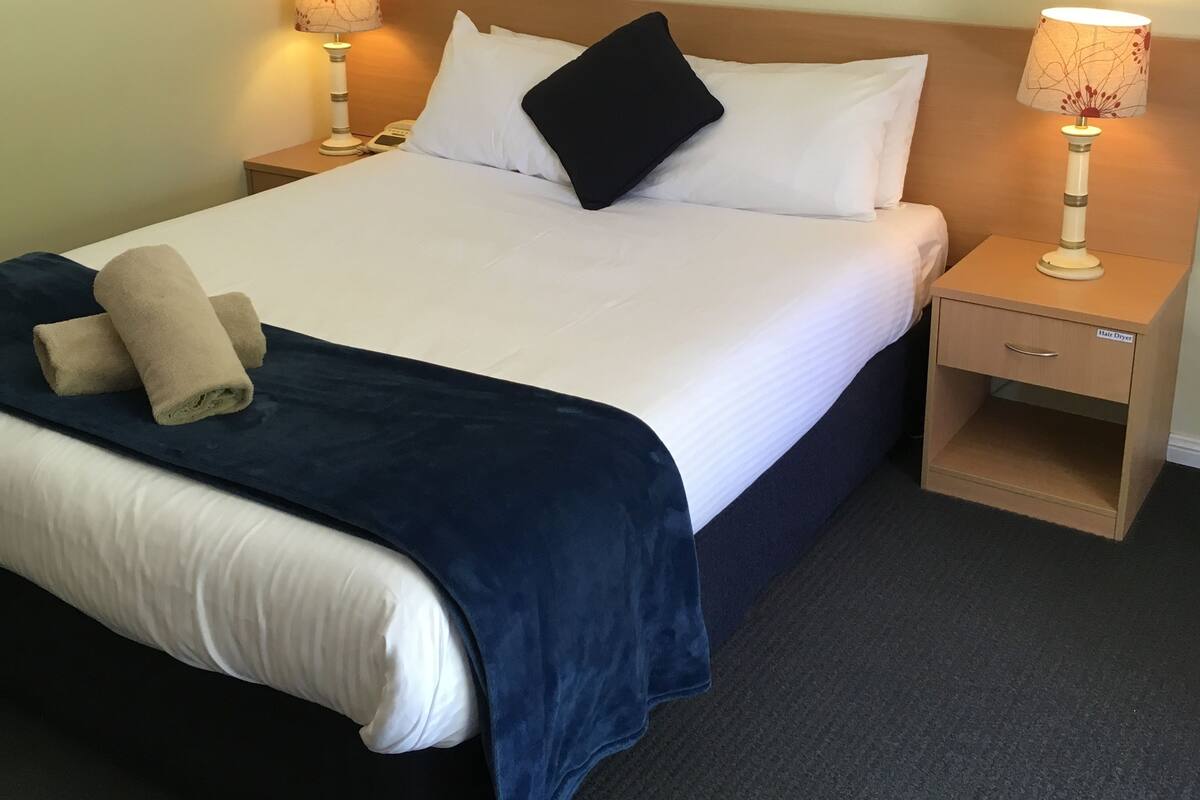 A neatly made bed with white linens, a dark blue bedspread, and a beige towel. There are two bedside tables with lamps, one of which has a label reading "Hair Dryer".