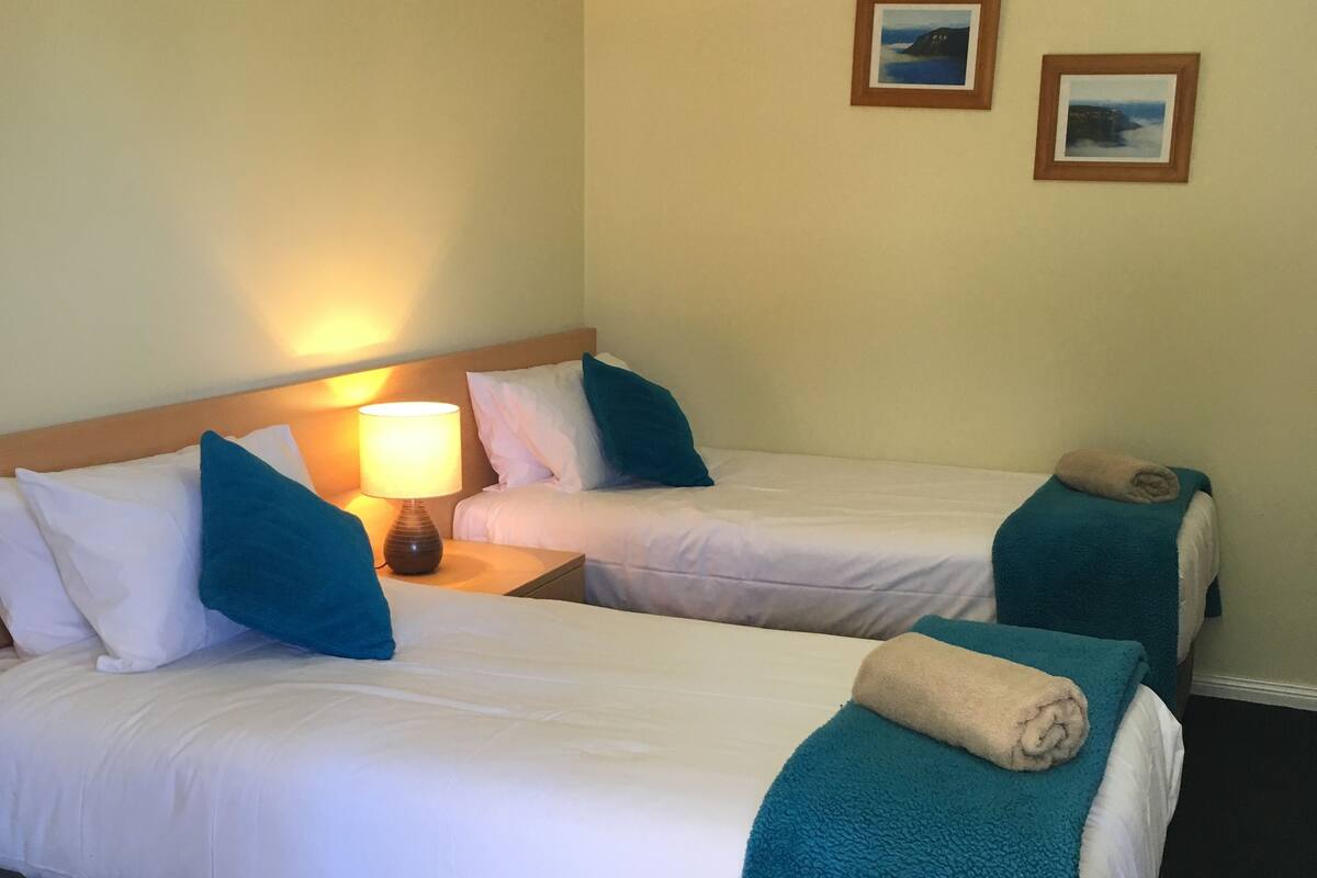 A hotel room with two beds, each with white bedding and teal throw pillows. There is a nightstand with a lamp between the beds.