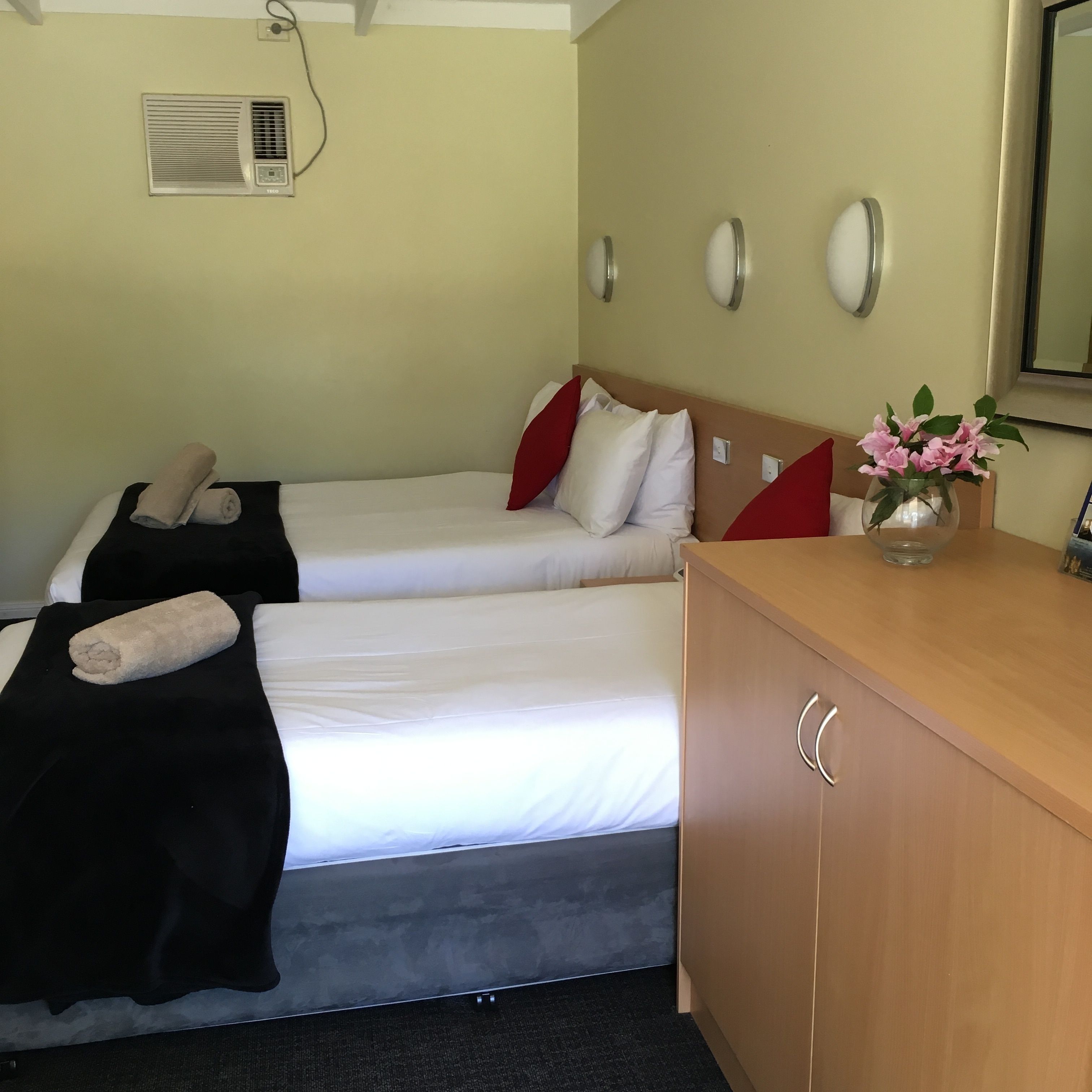 Une chambre d’hôtel avec deux lits, une commode en bois, un miroir et une bouche d’aération fixée au mur.