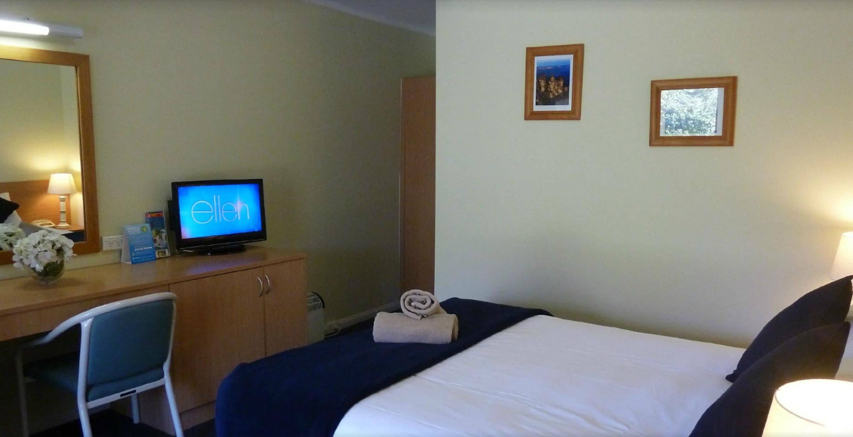 A hotel room with a bed, a desk with a computer, a chair, and two framed pictures on the wall.