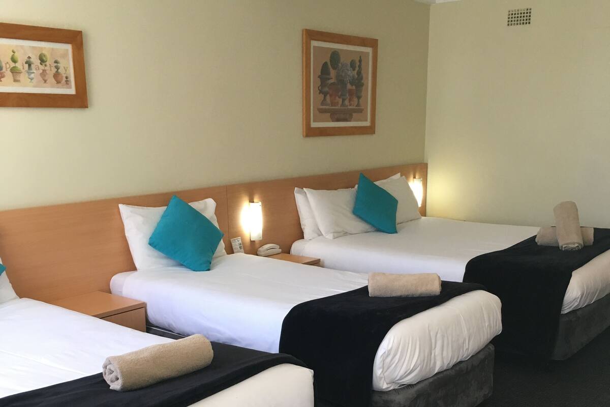 A hotel room with three beds, each with a black bedspread and white pillows. There is a wooden headboard, a wall-mounted phone, and a small table with a lamp.