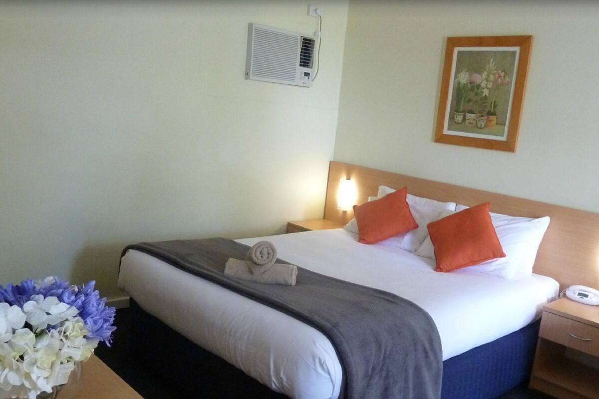 A hotel room with a bed, bedside table, lamp, and a floral arrangement.
