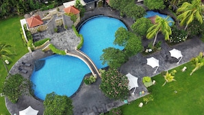 Uma ampla área de piscina ao ar livre, com várias piscinas, cercada por vegetação exuberante e palmeiras.
