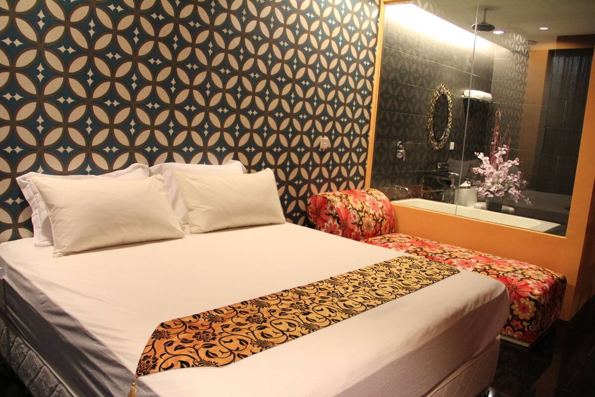 A hotel room with a large bed, a floral-patterned chair, a mirror, and a patterned wall.