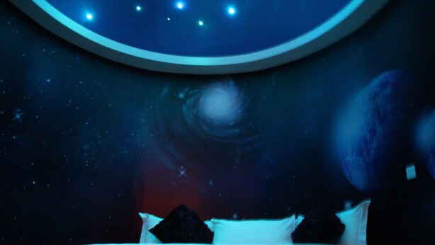 Quarto com cama, detalhe circular no teto e um mural na parede retratando uma cena espacial.