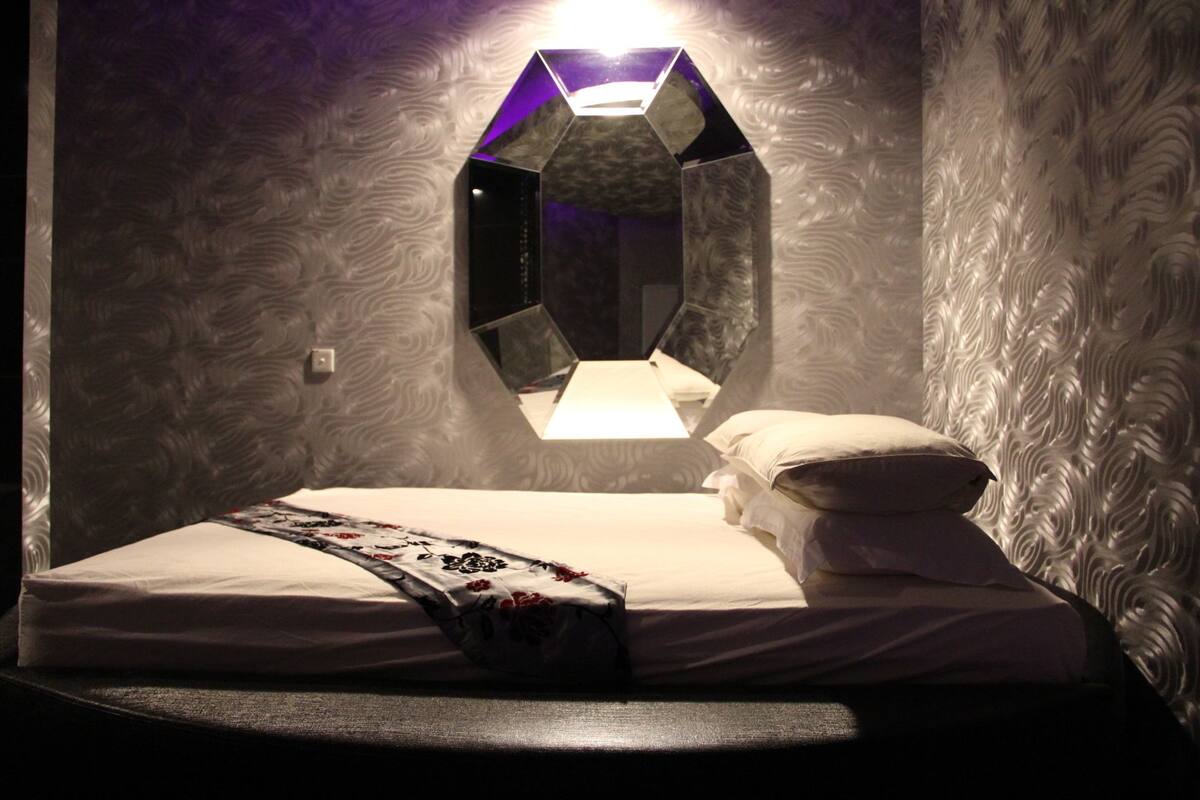 A room with a bed, a unique hexagonal mirror, and patterned wallpaper.