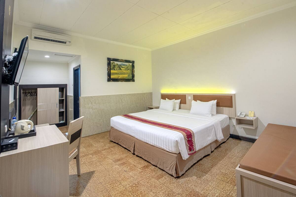 A hotel room with a bed, bedside table, a desk with a chair, a mirror, and a small cabinet.