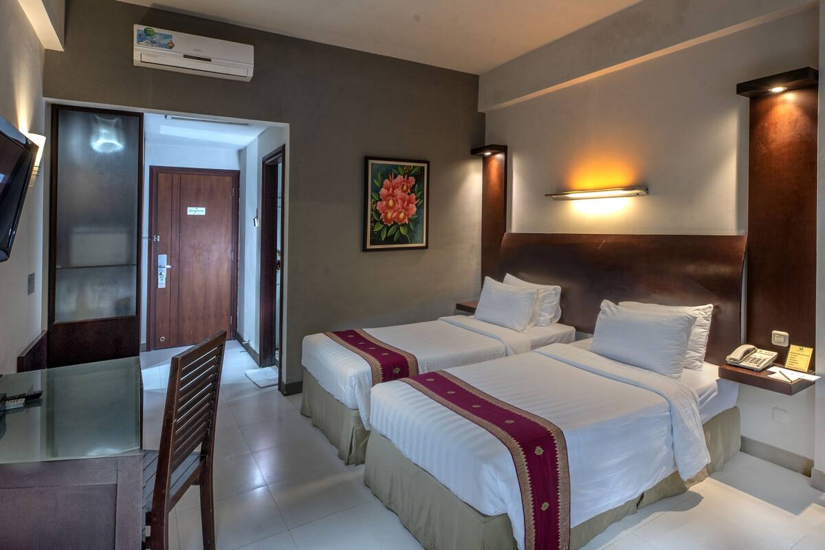 A hotel room with two beds, a desk with a chair, a TV, a painting, and a bedside table with a phone and a notepad.