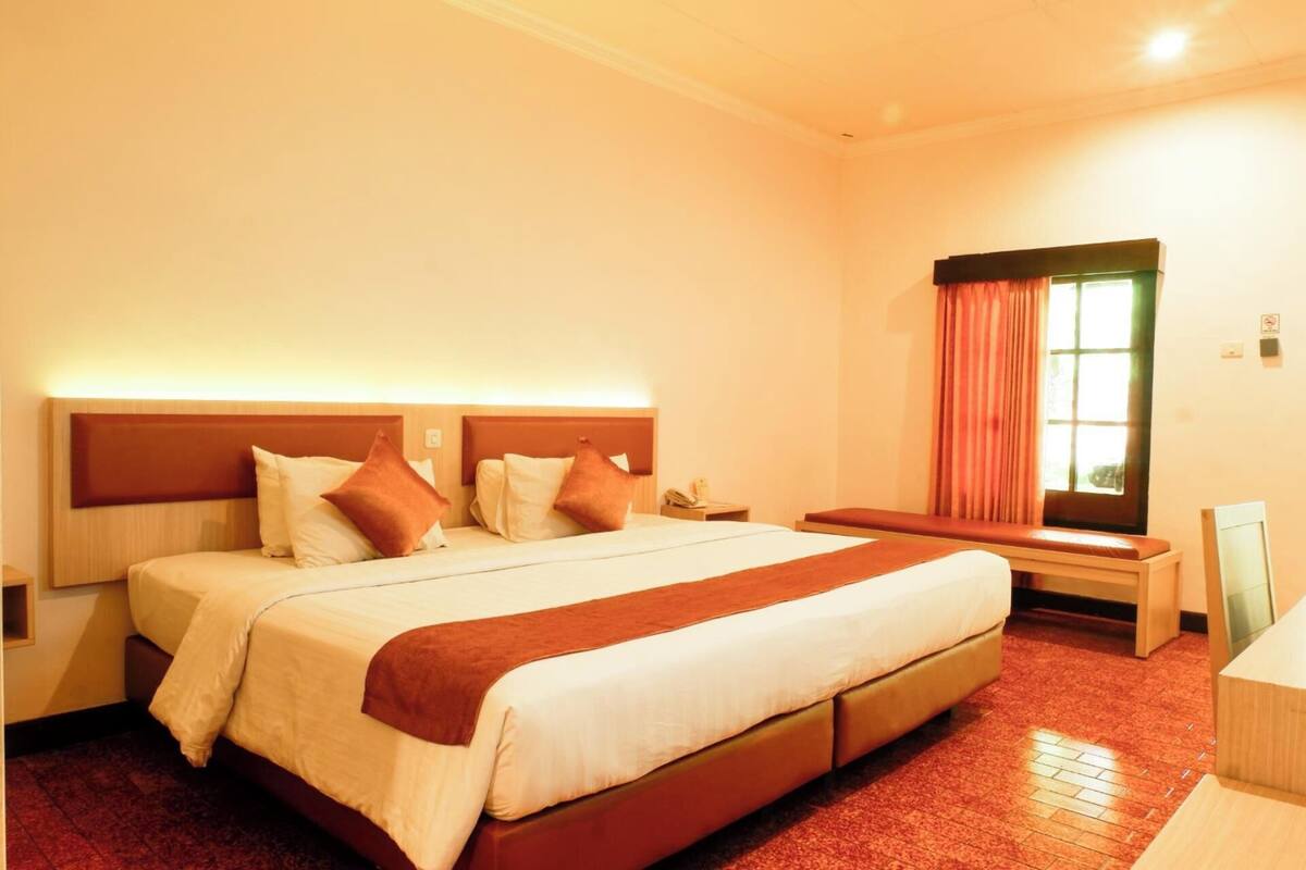 A hotel room with a large bed, a bedside table, a nightstand, a window with curtains, and a wall-mounted light.