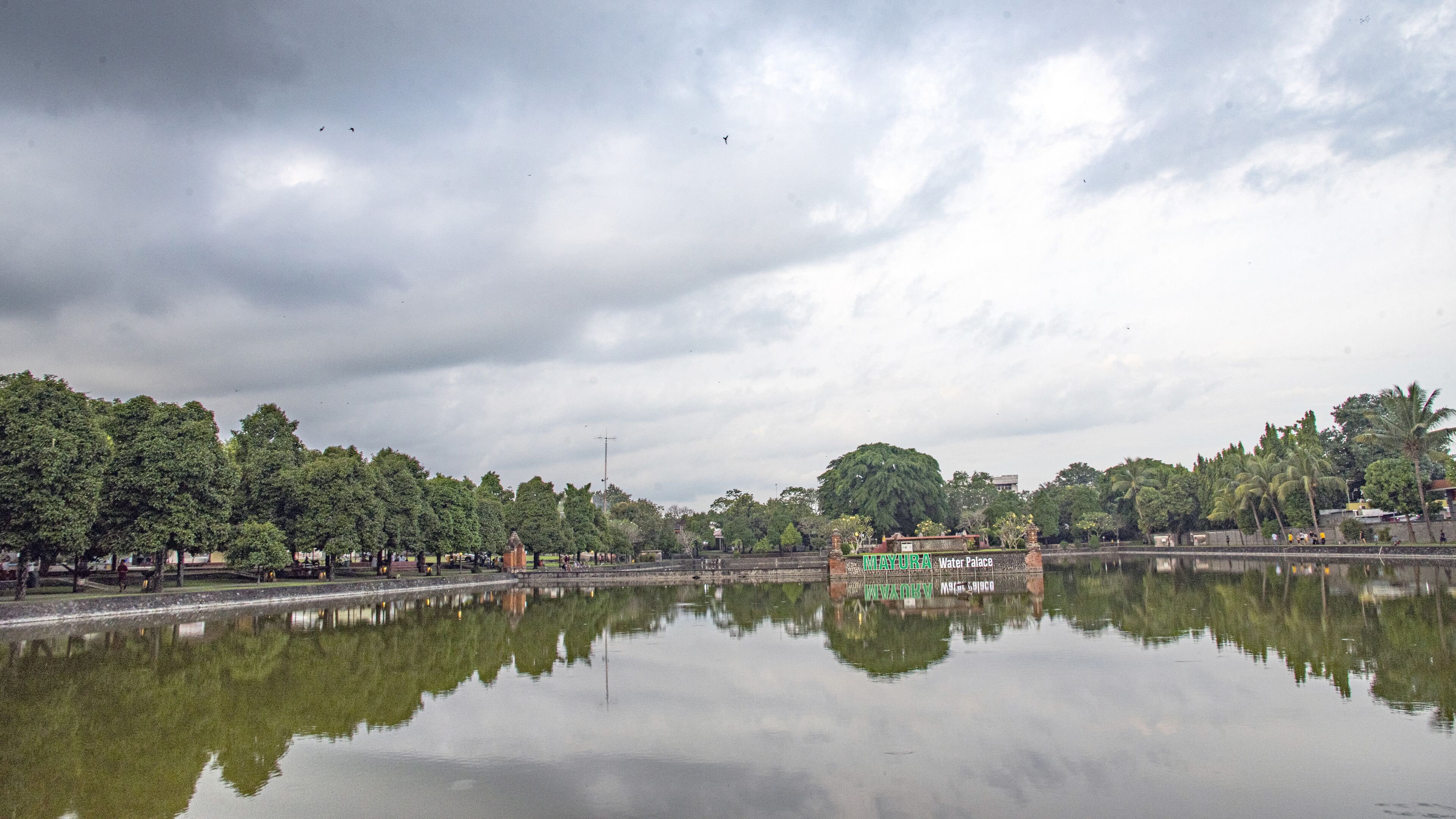 Un cuerpo de agua tranquila que refleja árboles y un edificio con la leyenda 'MAYURYA Water Palace'.
