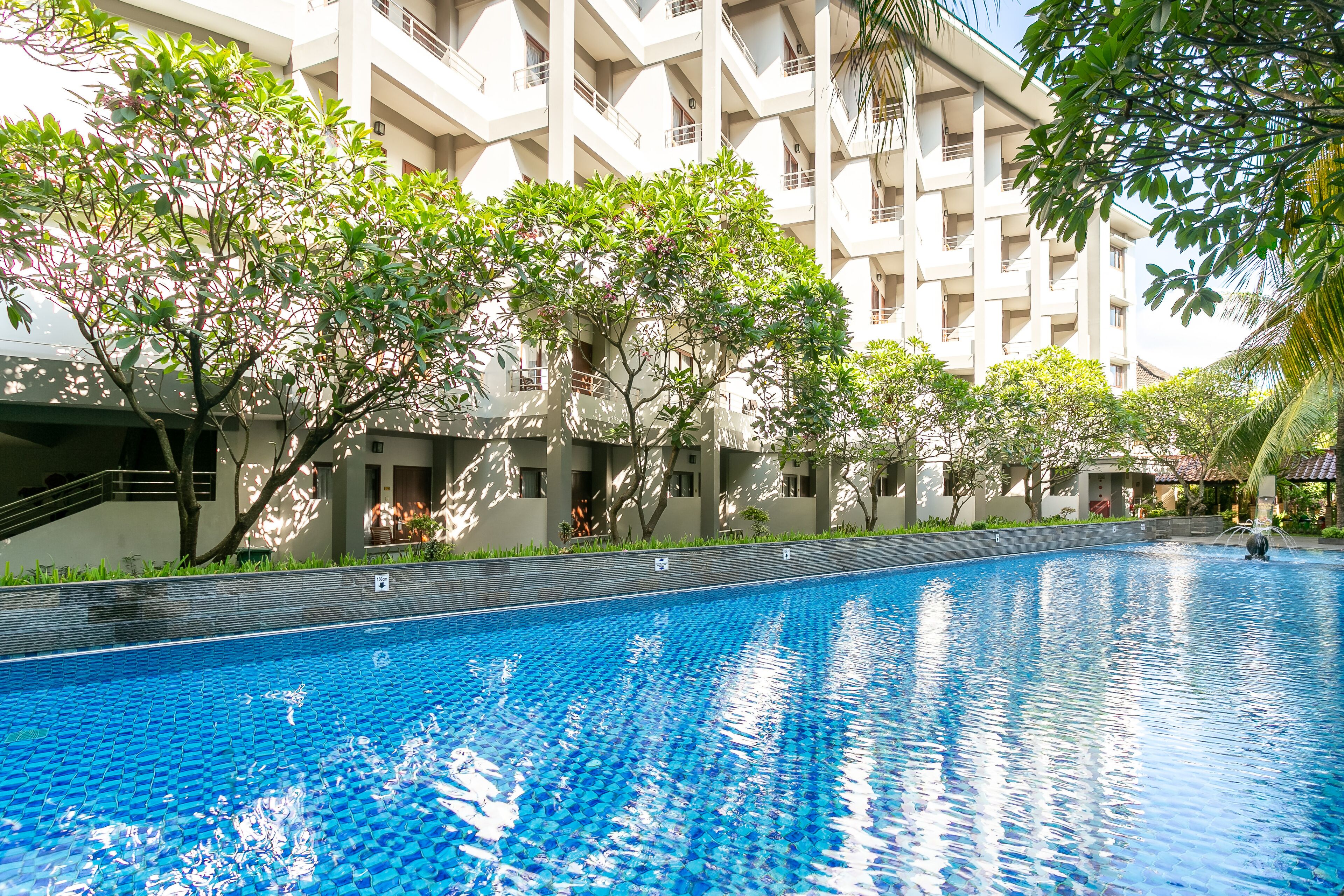 A swimming pool with a tiled edge, surrounded by trees and a multi-story building with balconies.