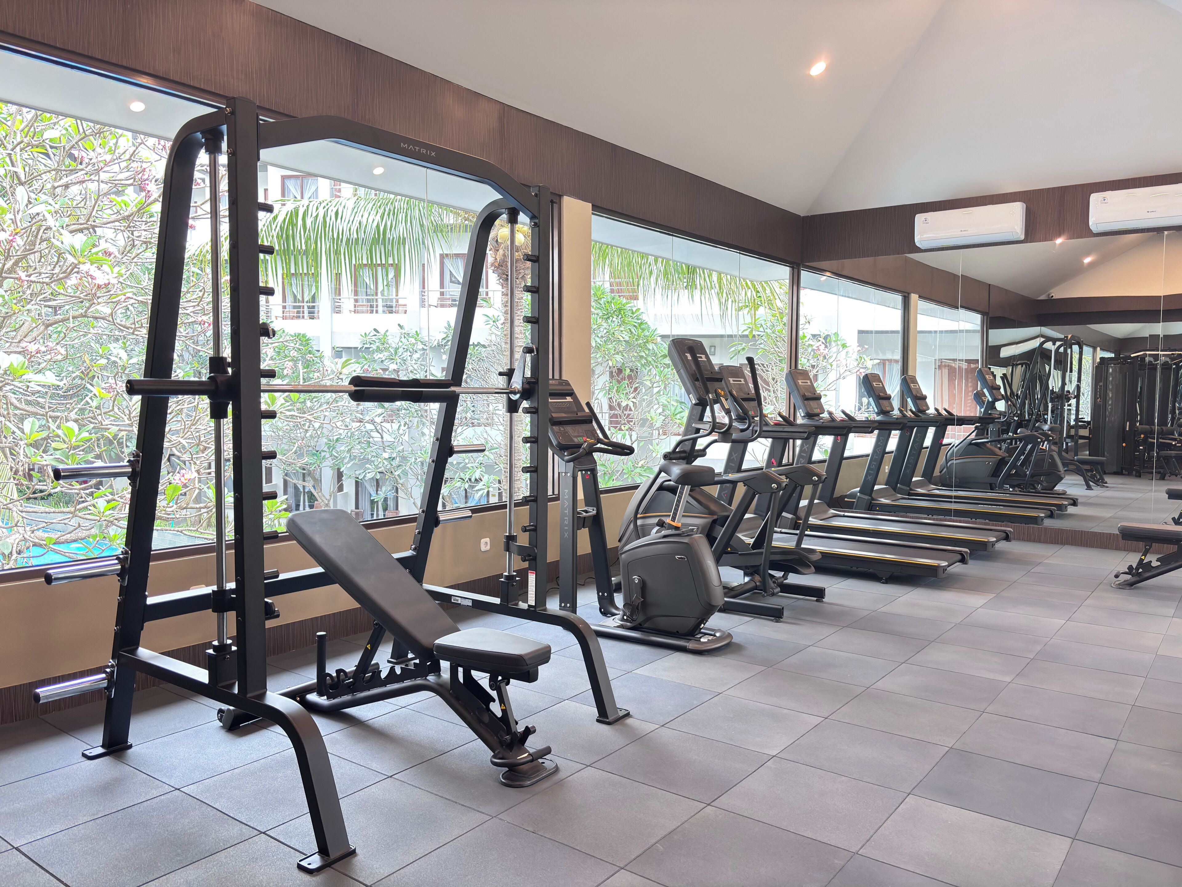 A gym with various exercise equipment, large windows, and a view of greenery outside.