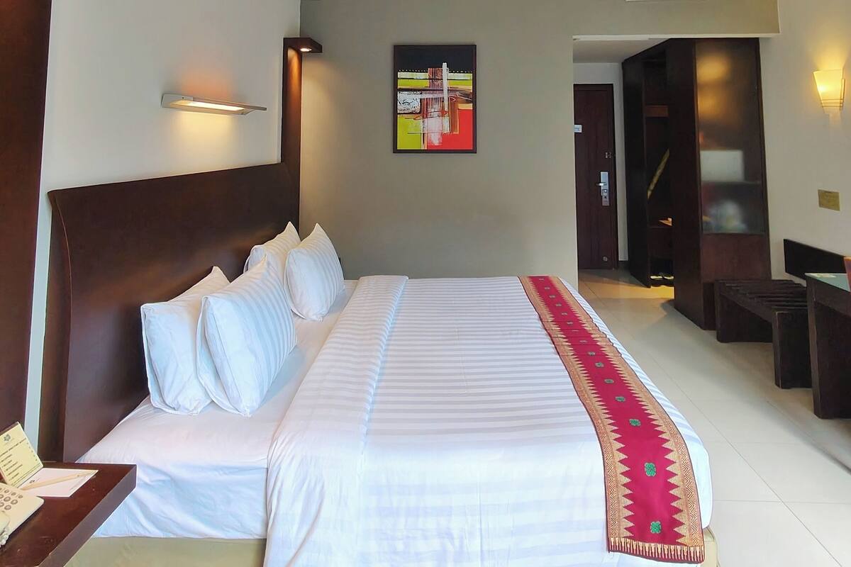 A hotel room with a large bed, a bedside table, a wall-mounted air conditioner, and a door leading to another room.