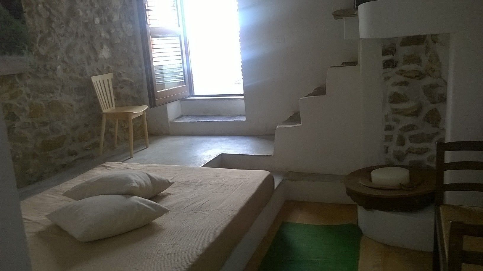 Un dormitorio con una cama, una silla, una mesita con una vela y una pared de piedra.