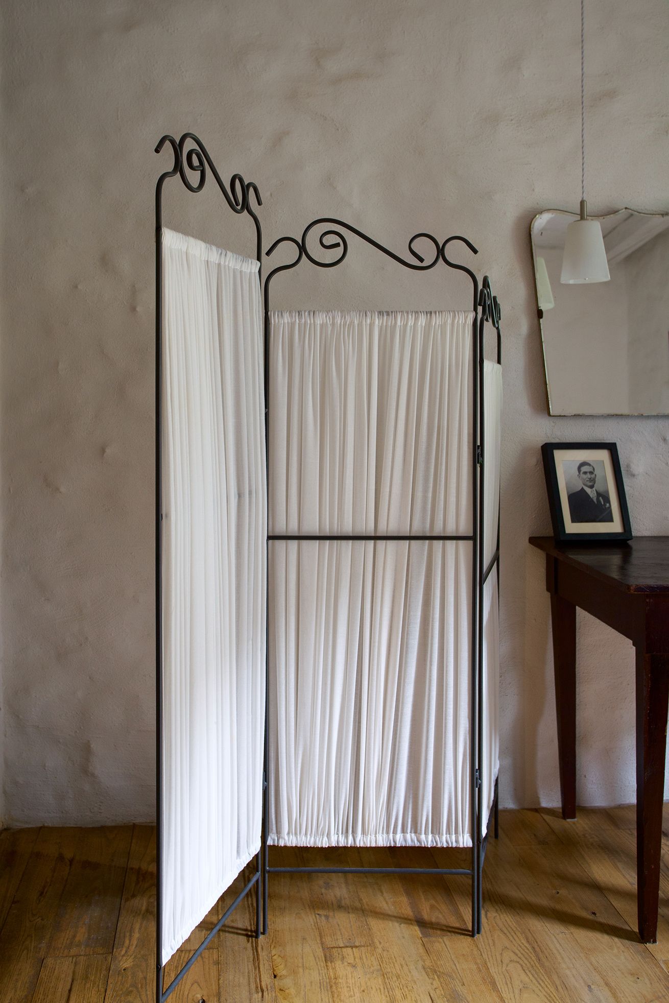 A room with a metal partition and white curtains, a wooden floor, a small table with a framed picture, and a hanging light fixture.