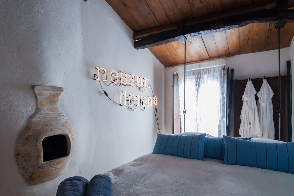 A bedroom with a bed, blue pillows, a rustic wall, a neon sign, and a hanging light fixture.