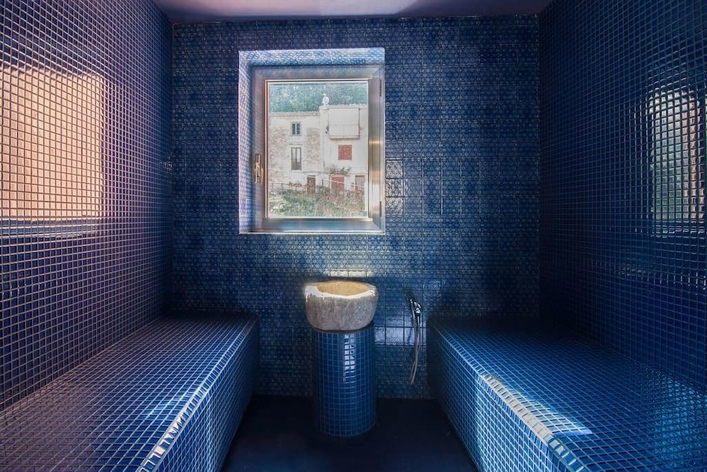 Ett badrum med blå mosaikplattor, ett fönster och ett handfat.