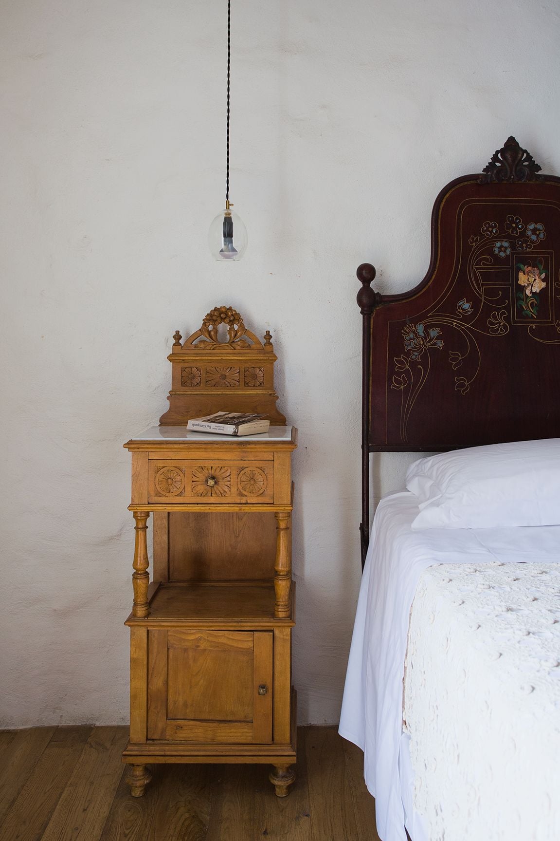 A wooden nightstand with a clock and a small lamp, a bed with a decorative headboard, and a plain wall.