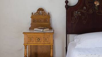 Una mesita de noche de madera con un reloj y una lámpara pequeña, una cama con cabecero decorativo y una pared sencilla.