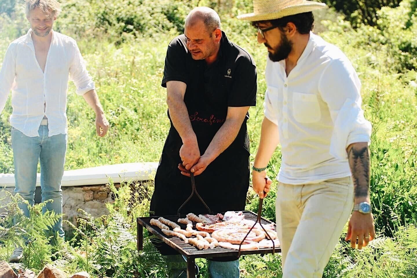 Three people grilling sausages outdoors with a stone wall and greenery in the background.