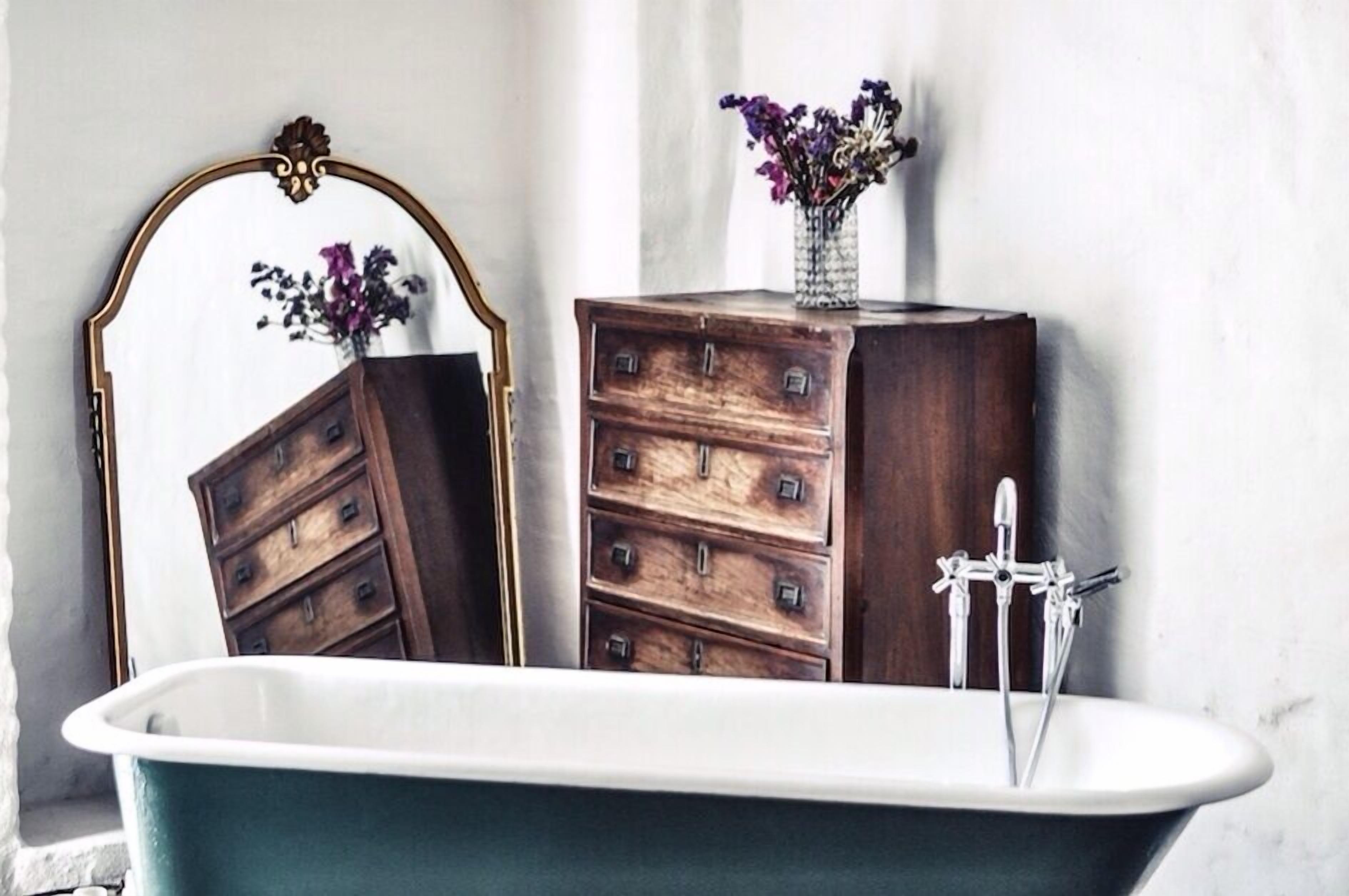 Un baño de estilo vintage con una bañera independiente, un aparador de madera con cajones, un espejo con un marco ornamentado y un jarrón con flores sobre el aparador.