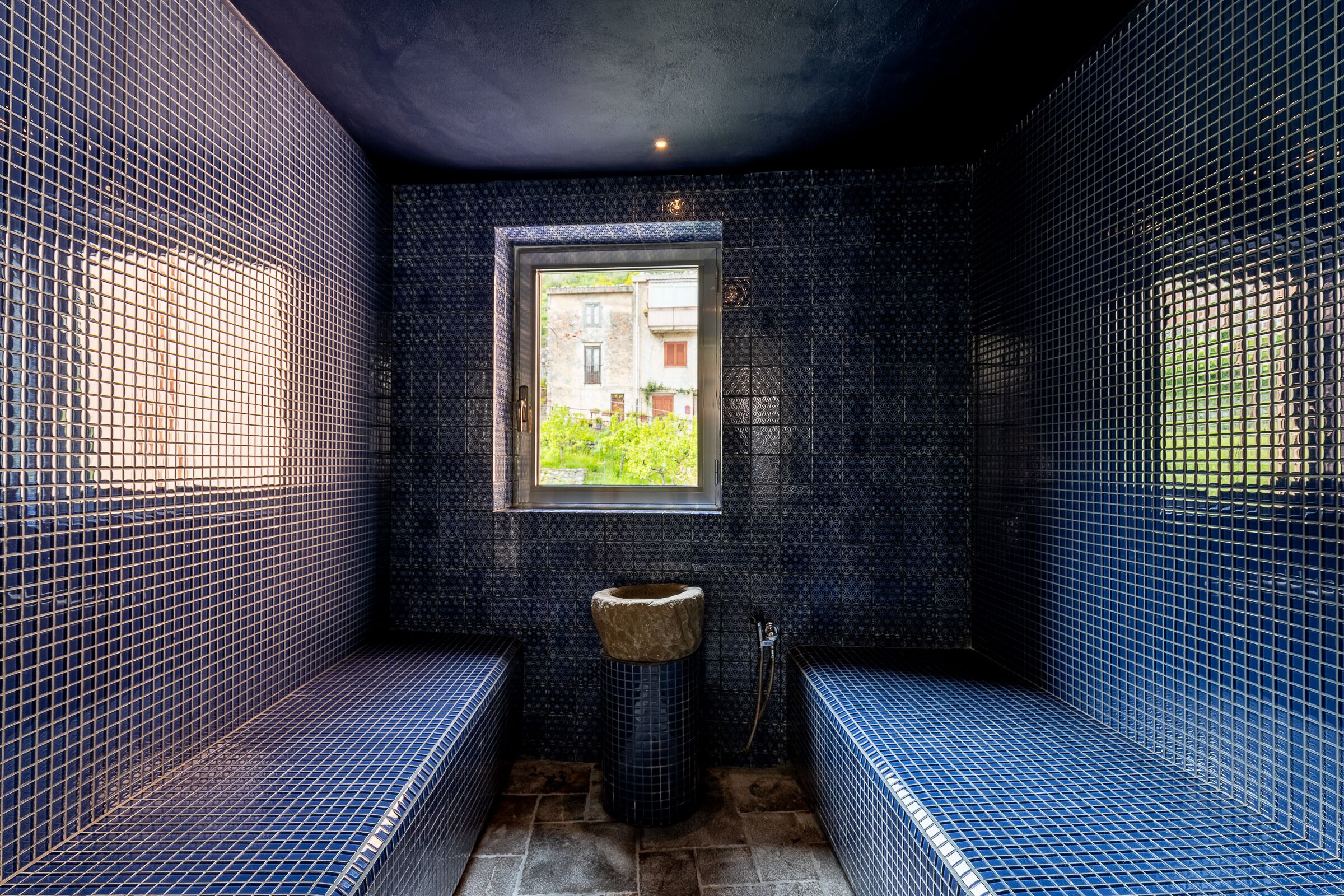 A bathroom with blue tiled walls, a window, a sink, and benches.