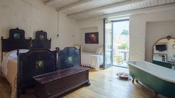 Un dormitorio con piso de madera, una cama doble, una mesita de noche, un aparador, una bañera verde y un balcón con vista a la ciudad.