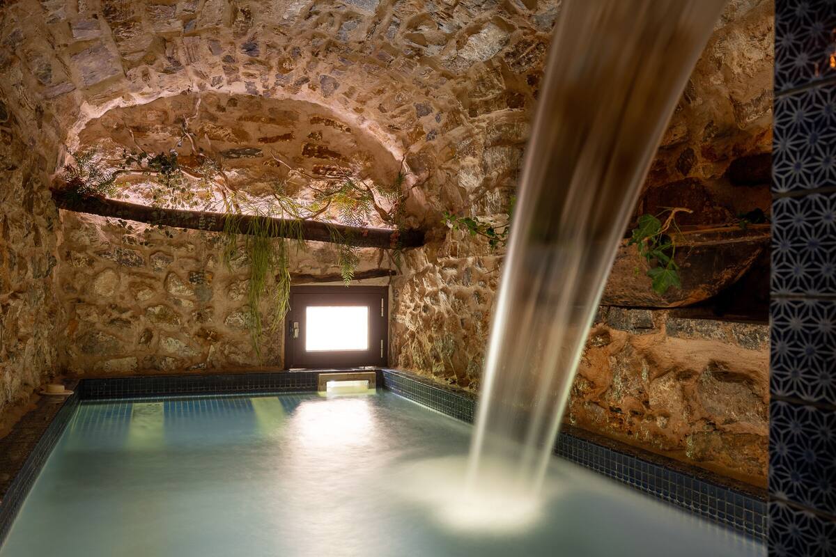 A stone-walled indoor pool with a waterfall feature, a bench, and a door leading outside.