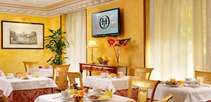 A hotel dining area with tables set for breakfast, a framed cityscape picture, a television displaying the hotel name, and a large plant.