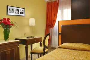 Una camera d'albergo con un letto, una scrivania, una sedia, una lampada, un vaso con fiori e un quadro appeso al muro.