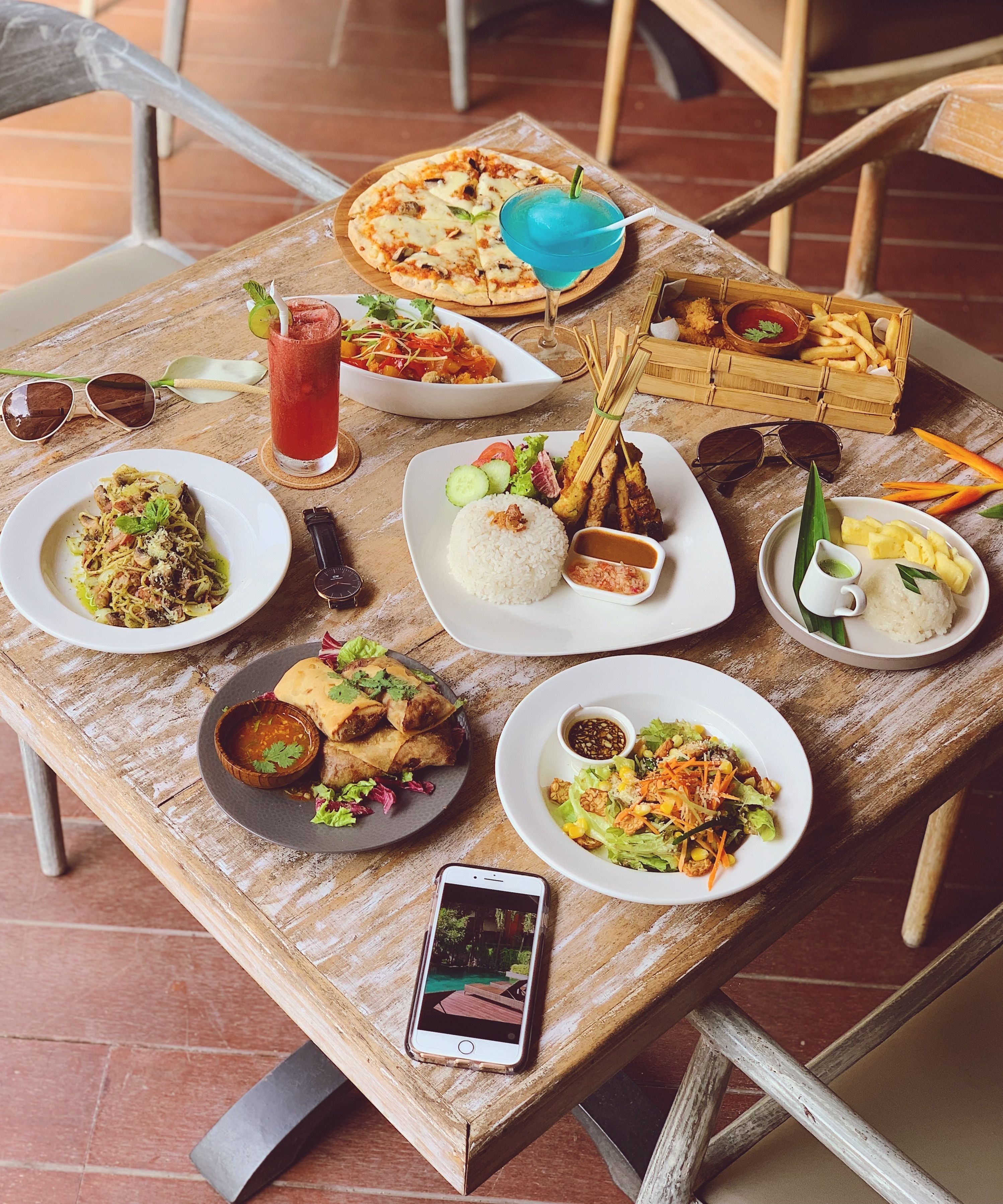 A wooden table with various dishes including pizza, rice, and salads, and a smartphone on it.