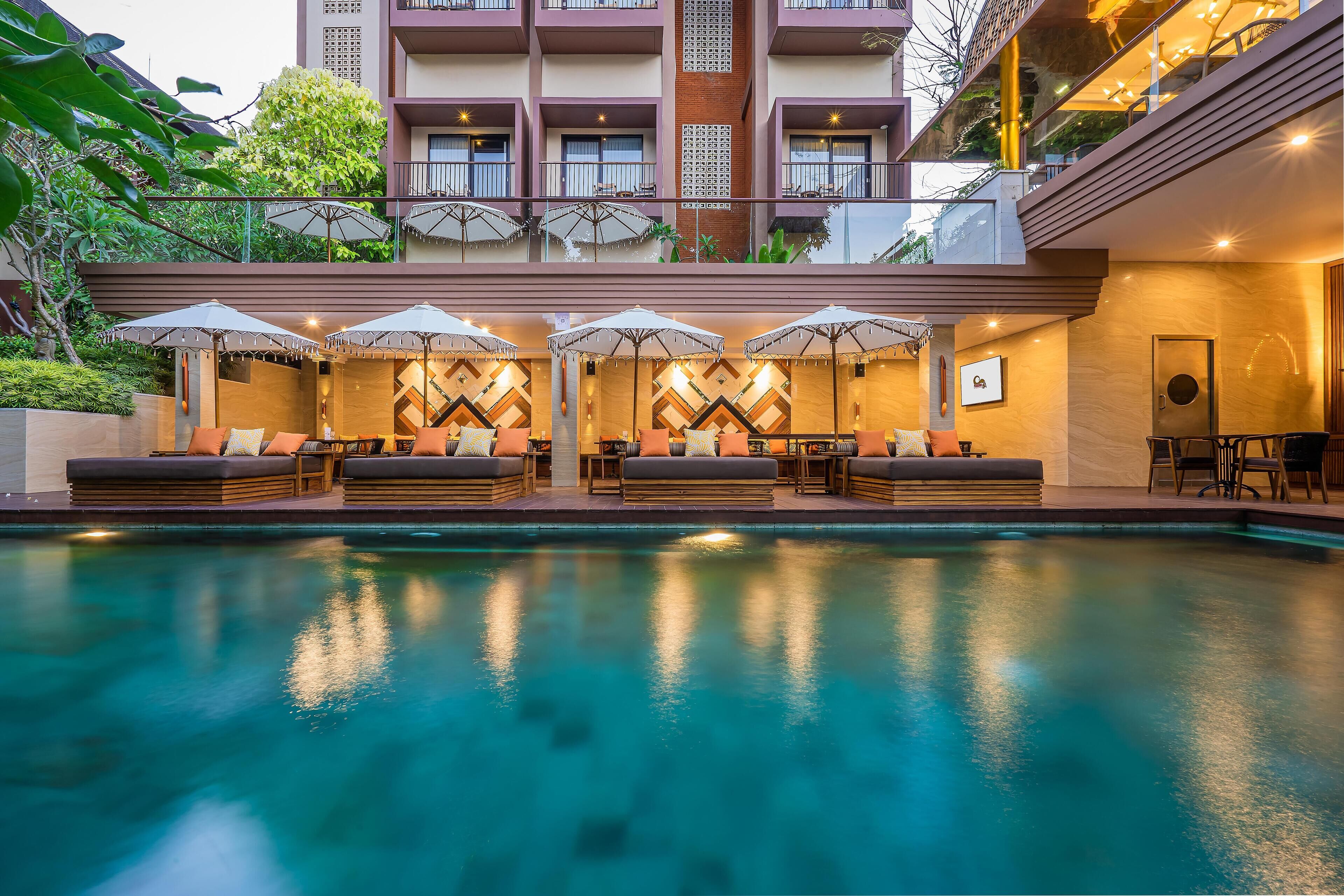 Zona de piscina con sillones de descanso y sombrillas, piscina y edificio con balcones y mesas al aire libre.