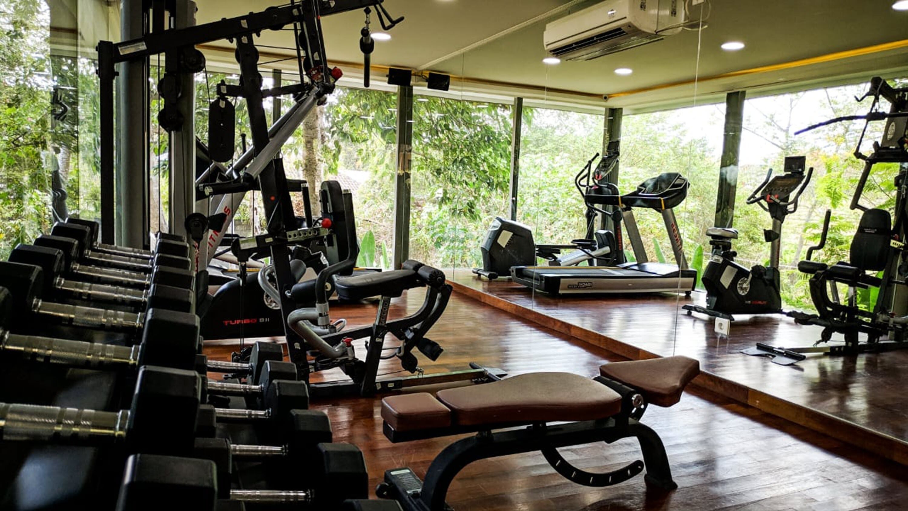 Un gimnasio con diversos equipos de ejercicio, amplias ventanas y suelo de madera.