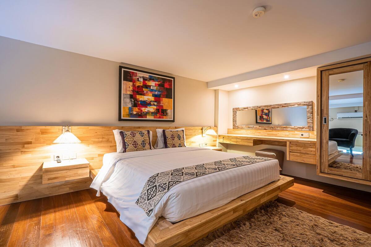 A bedroom with a wooden headboard, a large bed with patterned bedding, bedside lamps, a wooden nightstand, a mirror, and a colorful abstract painting on the wall.