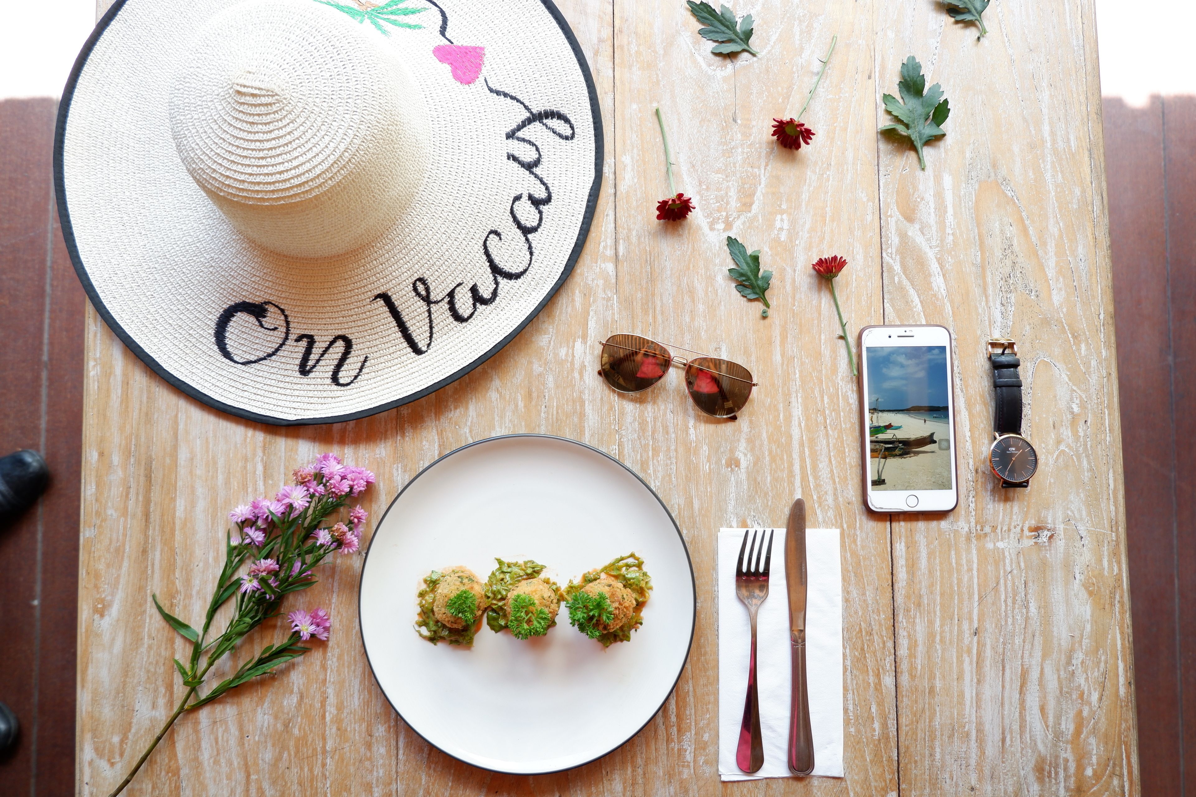 A dining table with a straw hat, a plate of food, a smartphone, a watch, and a fork and knife set.