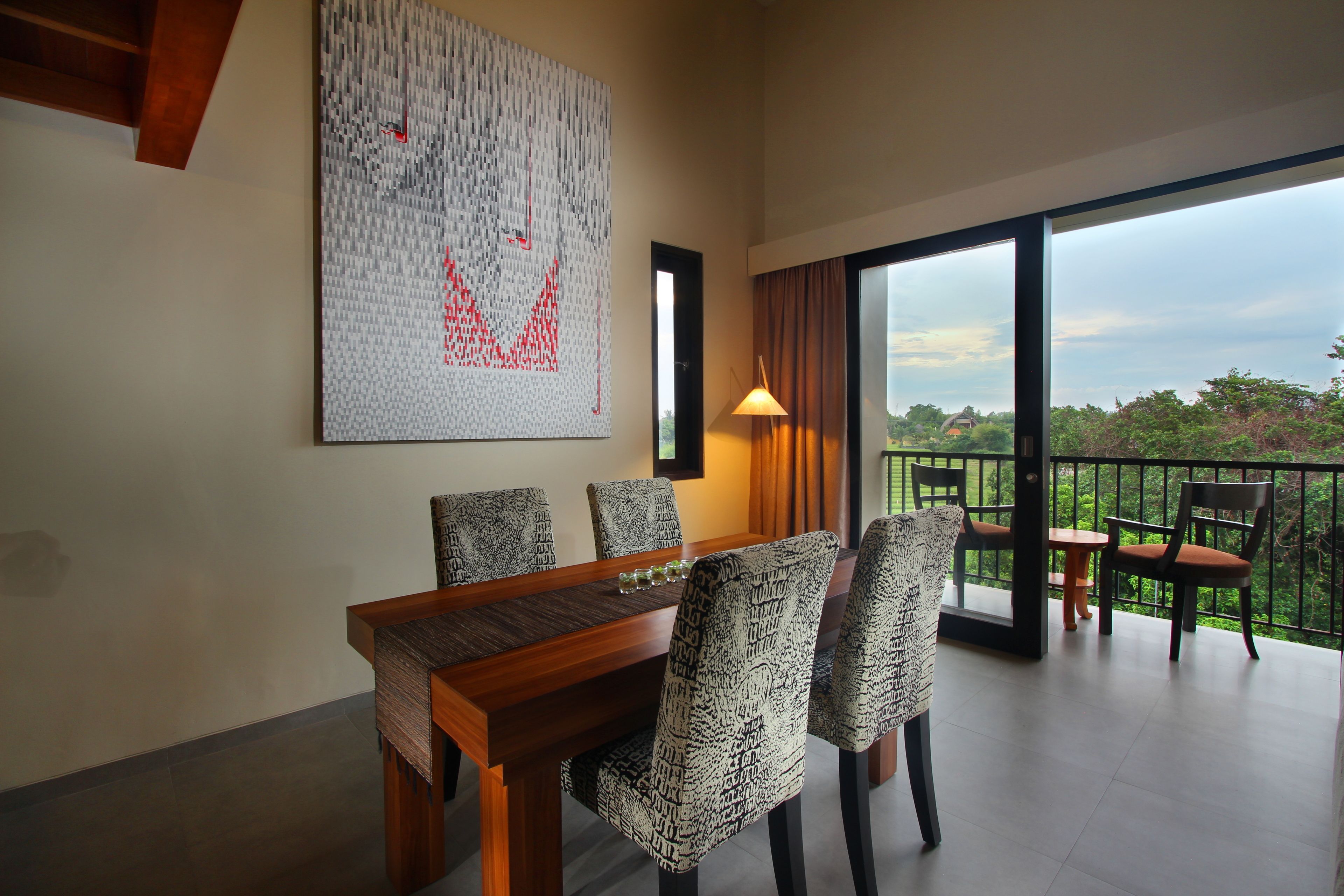 Un área de comedor con una mesa de madera y sillas estampadas, una gran obra de arte abstracta en la pared y un balcón con vistas a zonas verdes.