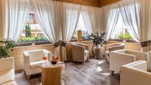 Una habitación con piso de madera, sillas tapizadas en blanco, una mesa de centro de madera y ventanas grandes con cortinas translúcidas.