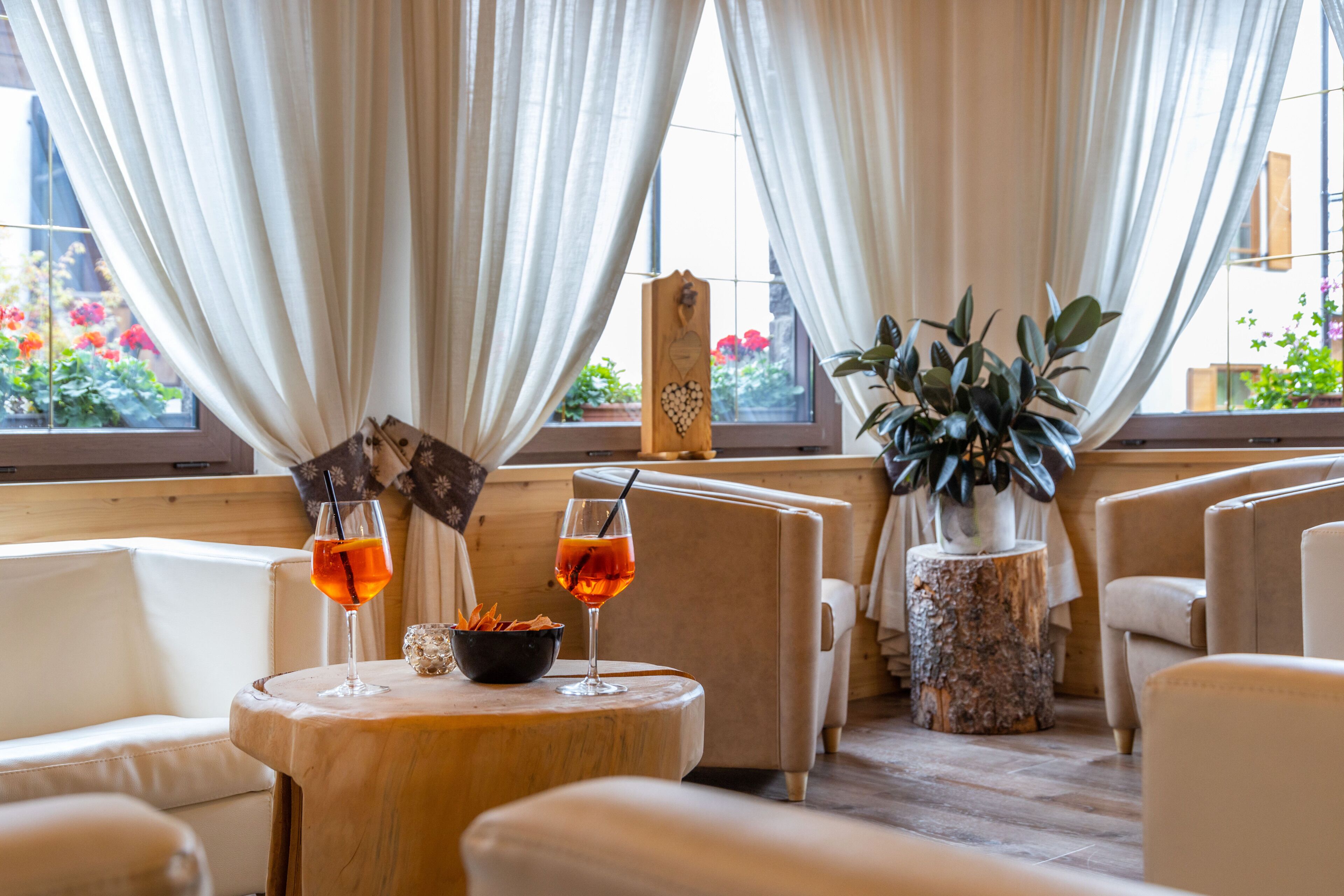 A cozy seating area with two round tables, each with a glass of orange drink and a bowl of snacks. The room has a wooden floor, white leather chairs, and a large window with curtains.