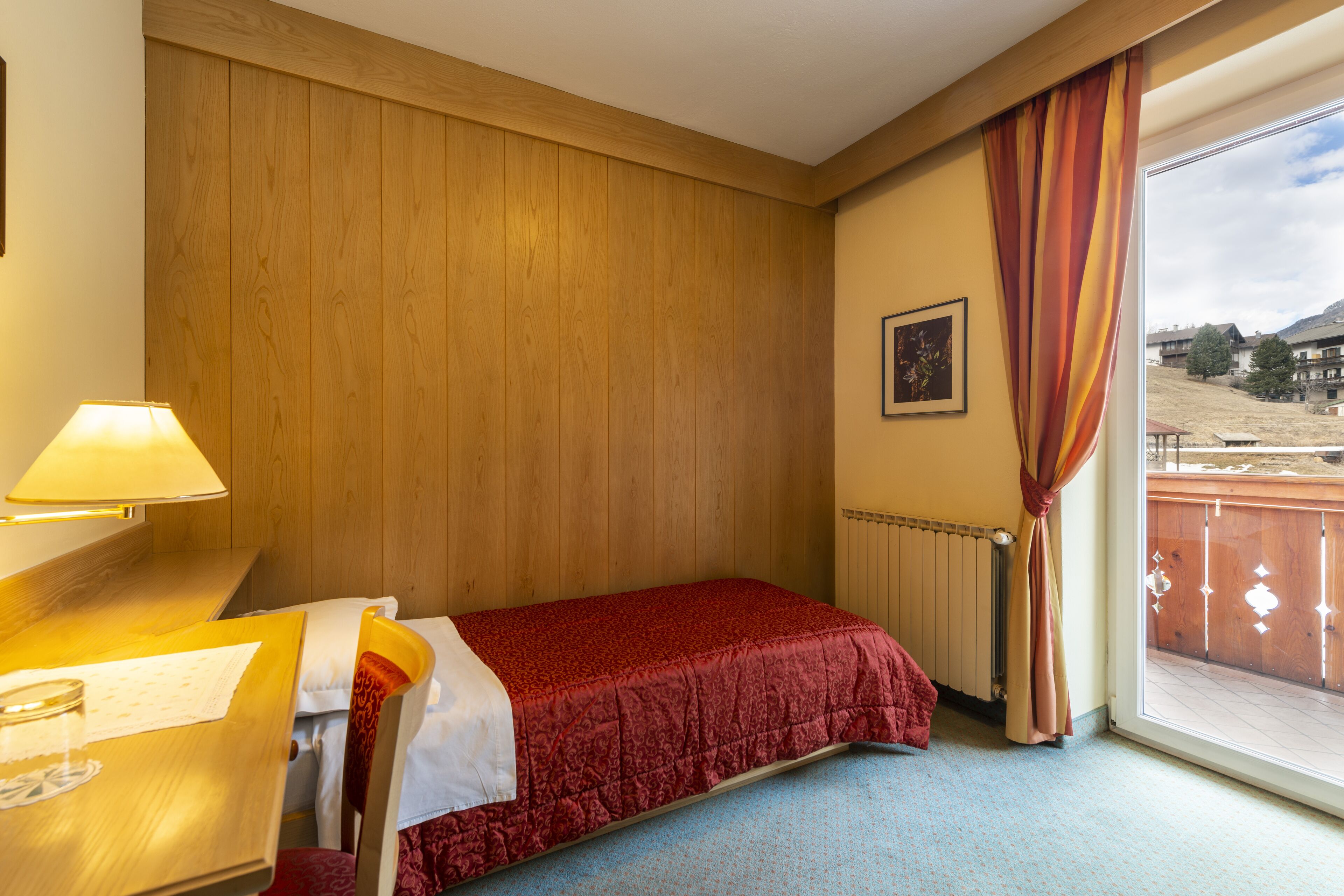 Une chambre d’hôtel avec un mur lambrissé en bois, un lit recouvert d’une courtepointe rouge, une fenêtre avec des rideaux et une vue sur un paysage montagneux.