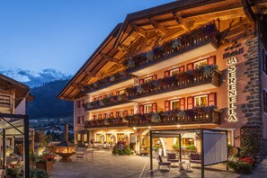 A hotel with a traditional alpine architectural style, featuring balconies with flower boxes, a well-lit entrance, and a courtyard with outdoor seating.
