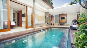 A modern pool area with a clear pool, wooden deck, and a seating area with a sofa and TV. - Samaja Villas Kunti (Seminyak)