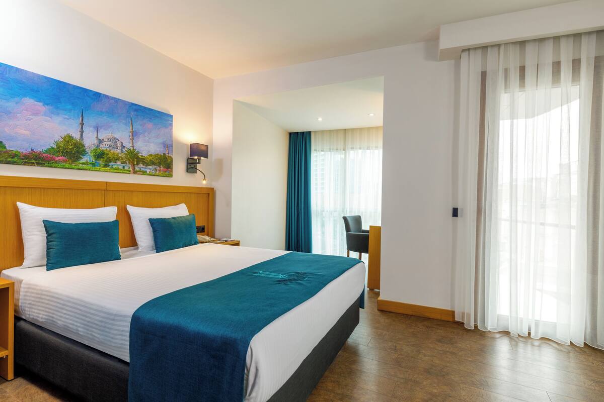 A hotel room with a large bed, a nightstand, a desk, a chair, a painting on the wall, and a view of the outside through the window.