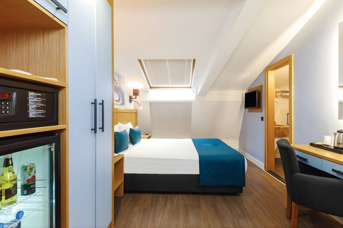 A compact hotel room with a bed, a desk, a chair, a mini-fridge, and a TV.