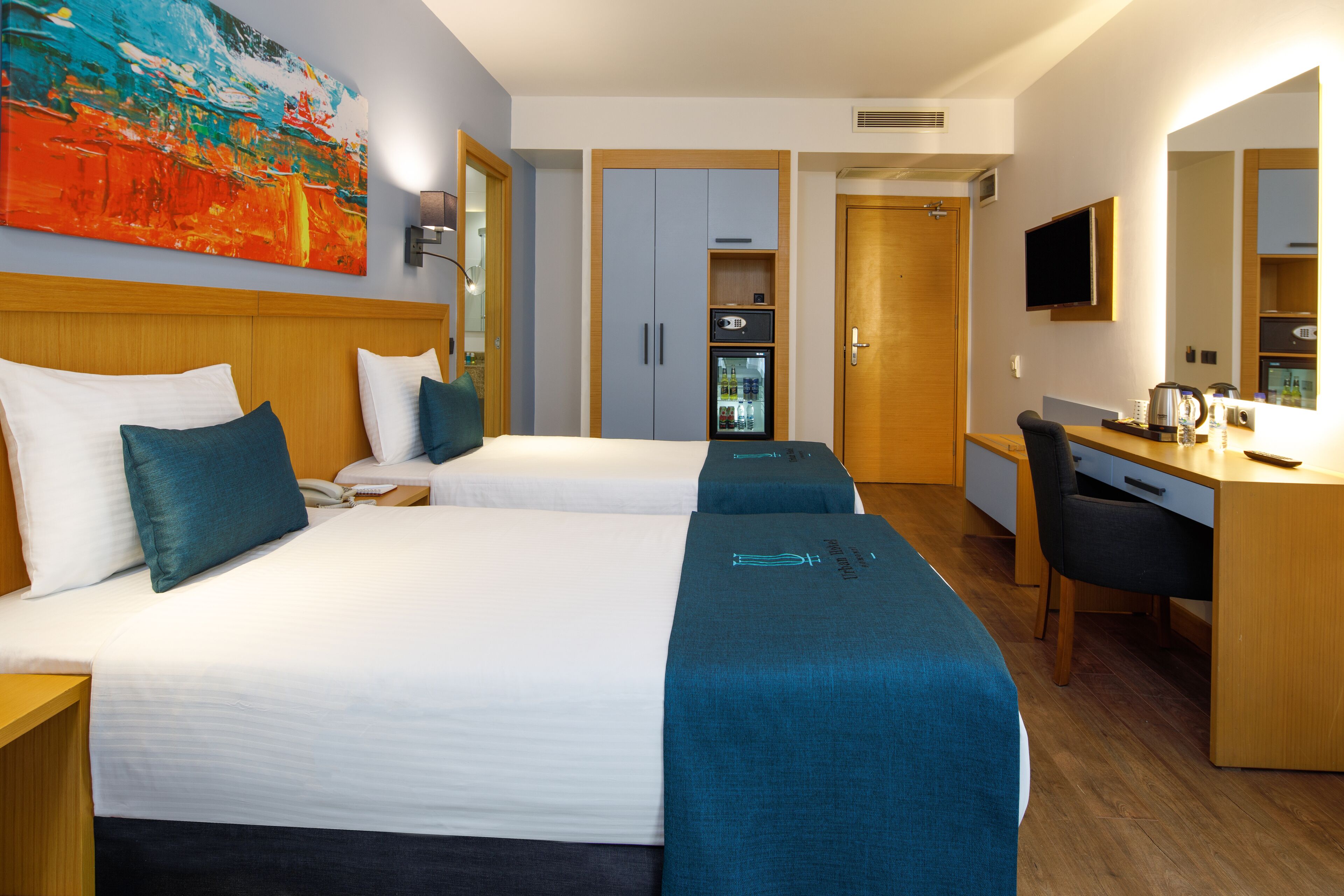 A hotel room with two beds, a desk, a chair, a refrigerator, and a TV.