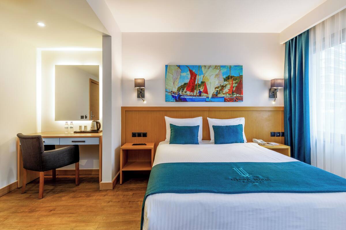 A hotel room with a bed, a desk, a chair, a painting on the wall, and a window with curtains.