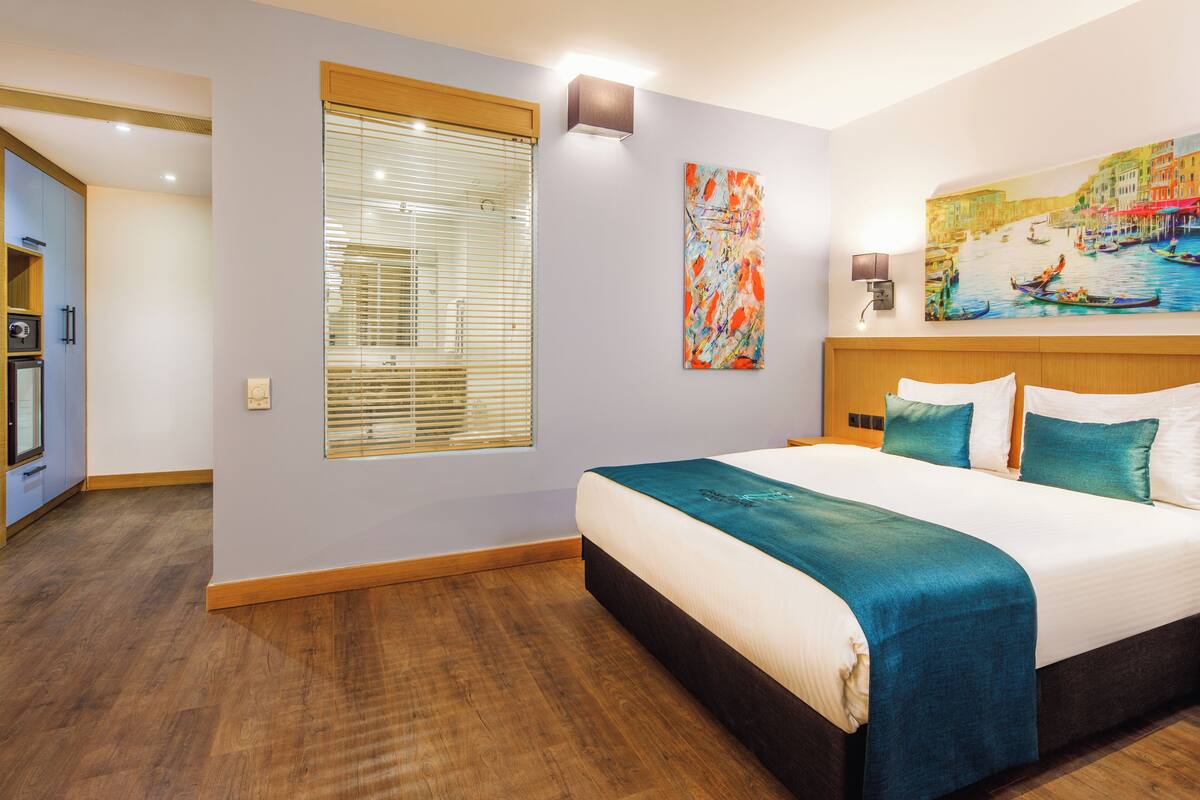 A hotel room with a large bed, a painting of a canal scene, a window with blinds, and a kitchenette area.