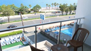 Ein Balkon mit zwei Holzstühlen und einem kleinen Tisch, der auf einen Poolbereich mit Liegestühlen und Sonnenschirmen, eine Straße mit Autos und ein Hotelschild blickt.